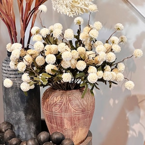 May include: A rustic, terracotta-colored vase with carved leaf patterns holds a bouquet of cream-colored, spherical flowers. A dark gray vase and a wooden tray with black beads are also visible, creating a warm, inviting home decor scene.