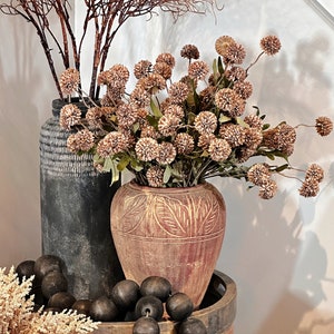 May include: A decorative arrangement featuring dried flowers and foliage in textured vases. The display includes a dark gray vase, a reddish-brown vase with carved details, and a wooden tray with dark wooden beads. The overall color palette is neutral with earthy tones.