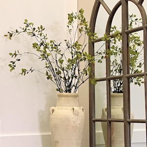 Peut inclure: Un grand vase en céramique blanc cassé avec deux poignées, rempli de branches vertes feuillues. Le vase est posé sur une petite table ronde en métal. Un grand miroir en forme de fenêtre arquée est en arrière-plan.