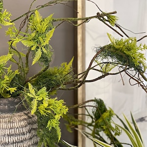 Peut inclure: Gros plan sur une composition décorative avec des feuilles vertes vives en forme de fougère et de la mousse, associées à des branches brunes. L'arrangement est placé dans un vase gris foncé texturé. Le style est naturel et botanique.
