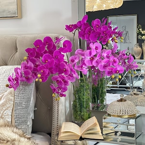 Peut inclure: Une table de chevet avec un jeté en fausse fourrure, un livre et un tiroir miroir avec un bouton en cristal. Un vase de fleurs d'orchidées roses est posé sur la table.