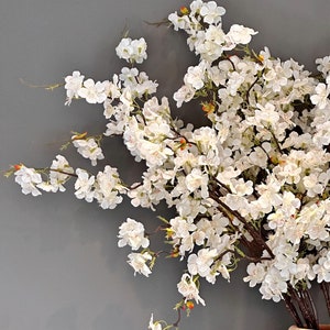 May include: A bouquet of white cherry blossoms arranged in a ceramic vase. The blossoms are delicate and have a soft, feathery texture. The vase is a light brown color and has a textured surface.