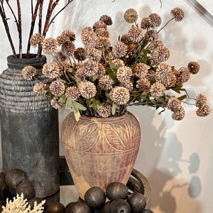 May include: An arrangement of decorative items featuring two textured vases, one dark gray and the other reddish-brown with a leaf pattern. The vases hold brown branches and dried flowers. Dark wooden beads and a small plant are in the foreground.