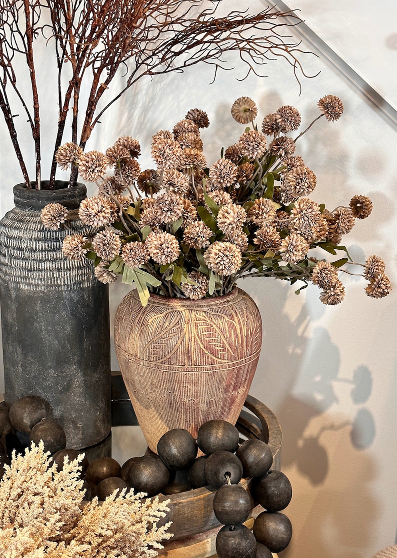 May include: An arrangement of decorative items featuring two textured vases, one dark gray and the other reddish-brown with a leaf pattern. The vases hold brown branches and dried flowers. Dark wooden beads and a small plant are in the foreground.