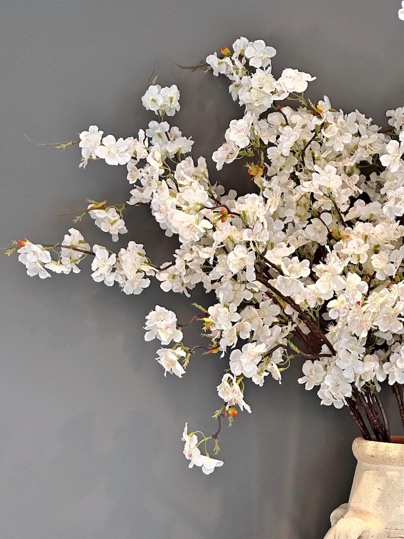 May include: A bouquet of white cherry blossoms arranged in a ceramic vase. The blossoms are delicate and have a soft, feathery texture. The vase is a light brown color and has a textured surface.