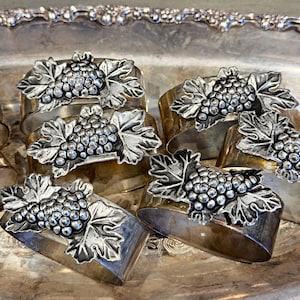 May include: A set of metallic napkin rings, each featuring a raised, silver-toned grape cluster and leaf motif attached to a smooth, curved band. The finish on the bands appears to have a warm, brassy or tarnished patina, contrasting with the bright, textured silver-toned grape embellishments.