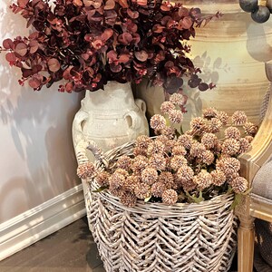 May include: A decorative arrangement featuring burgundy eucalyptus branches in a cream-colored vase, and a woven basket filled with tan globe-shaped flowers. The basket has a chevron pattern and is placed on a dark surface.
