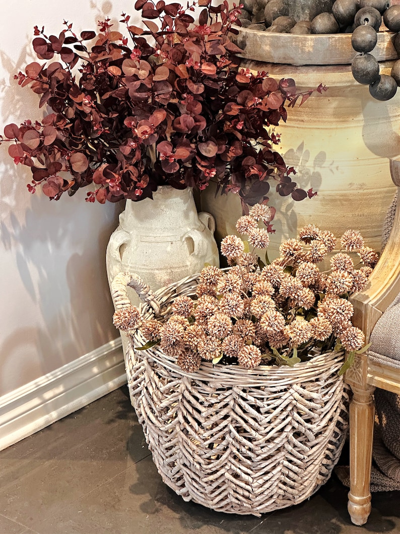May include: A decorative arrangement featuring burgundy eucalyptus branches in a cream-colored vase, and a woven basket filled with tan globe-shaped flowers. The basket has a chevron pattern and is placed on a dark surface.