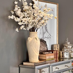 May include: A white ceramic vase with a textured surface and two handles sits on a silver mirrored dresser. The vase is filled with a bouquet of white cherry blossom branches. The dresser has two drawers and a mirrored surface. There are books stacked on the dresser, including "The Last Two Million Years".