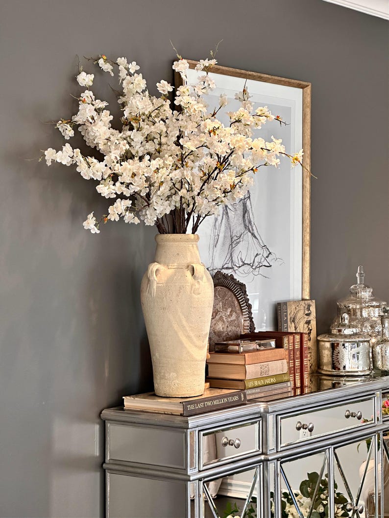 May include: A white ceramic vase with a textured surface and two handles sits on a silver mirrored dresser. The vase is filled with a bouquet of white cherry blossom branches. The dresser has two drawers and a mirrored surface. There are books stacked on the dresser, including "The Last Two Million Years".