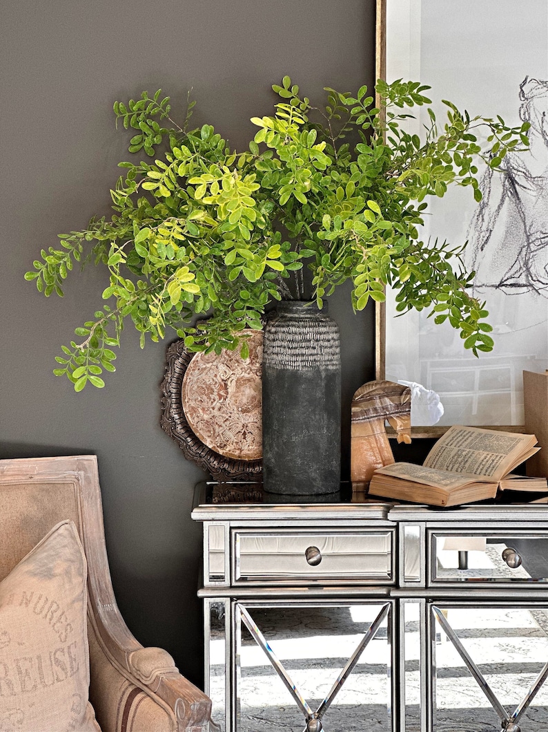May include: A dark gray wall with a vase of green leafy branches. The vase is black with a textured surface. The vase is sitting on a mirrored side table with two drawers. A book is on the table next to the vase.