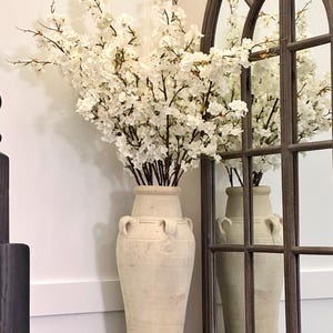 May include: A white ceramic vase with a textured surface and two handles on each side. The vase is filled with white cherry blossom branches.