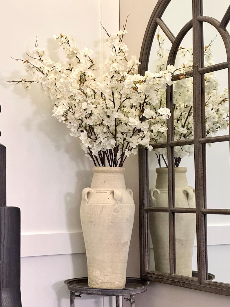 May include: A white ceramic vase with a textured surface and two handles on each side. The vase is filled with white cherry blossom branches.