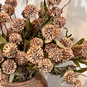 May include: A decorative arrangement of artificial flowers in a ceramic vase. The flowers are a mix of brown and tan, with green leaves. The vase is a reddish-brown color with carved details.