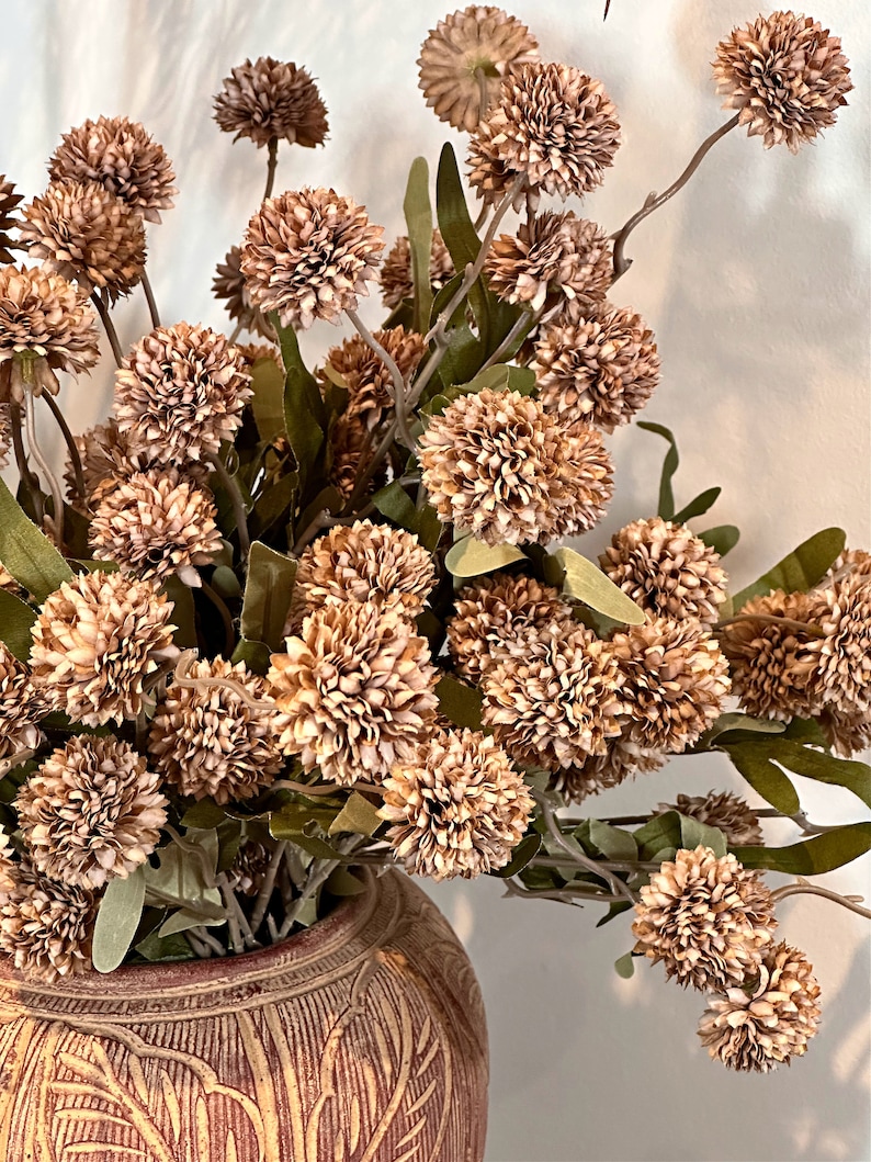 May include: A decorative arrangement of artificial flowers in a ceramic vase. The flowers are a mix of brown and tan, with green leaves. The vase is a reddish-brown color with carved details.