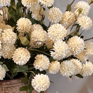 May include: A close-up of a floral arrangement featuring numerous cream-colored, spherical flowers with textured petals. The flowers are clustered on slender, brown stems with green leaves. The arrangement is in a decorative, textured brown vase.