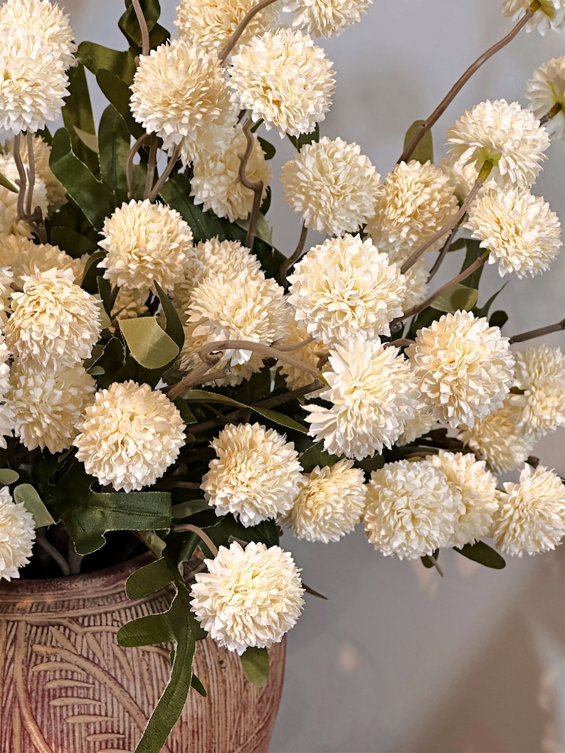 May include: A close-up of a floral arrangement featuring numerous cream-colored, spherical flowers with textured petals. The flowers are clustered on slender, brown stems with green leaves. The arrangement is in a decorative, textured brown vase.