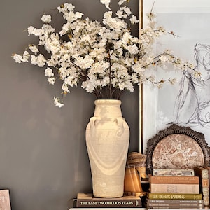 May include: A white ceramic vase with an ornate design, filled with a bouquet of white cherry blossoms. The vase is sitting on a mirrored table with a stack of books, including "The Last Two Million Years", "The Greatest Game of All", "Les Beaux Jardins", "Cage des Rois", and "Le Japon Medieval".
