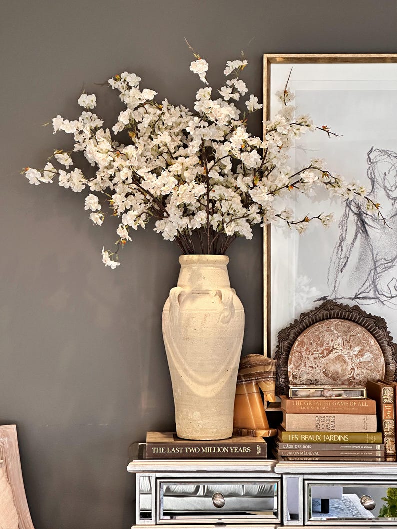 May include: A white ceramic vase with an ornate design, filled with a bouquet of white cherry blossoms. The vase is sitting on a mirrored table with a stack of books, including "The Last Two Million Years", "The Greatest Game of All", "Les Beaux Jardins", "Cage des Rois", and "Le Japon Medieval".