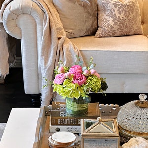 Op de afbeelding: Een witte bank met twee kussens, een met een bloemenpatroon en een effen kleur. Een dienblad op een salontafel bevat een vaas met roze en groene bloemen, een boek getiteld "Beautiful Chaos", een klein doosje en een decoratieve glazen stolp.