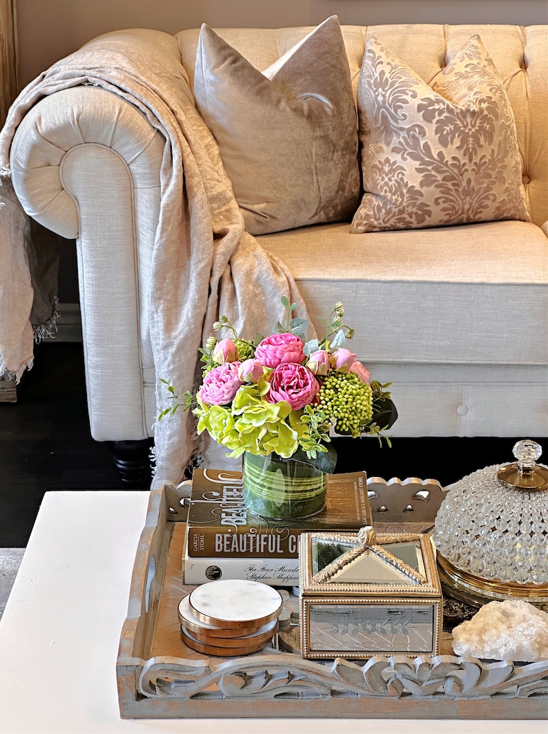 Op de afbeelding: Een witte bank met twee kussens, een met een bloemenpatroon en een effen kleur. Een dienblad op een salontafel bevat een vaas met roze en groene bloemen, een boek getiteld "Beautiful Chaos", een klein doosje en een decoratieve glazen stolp.