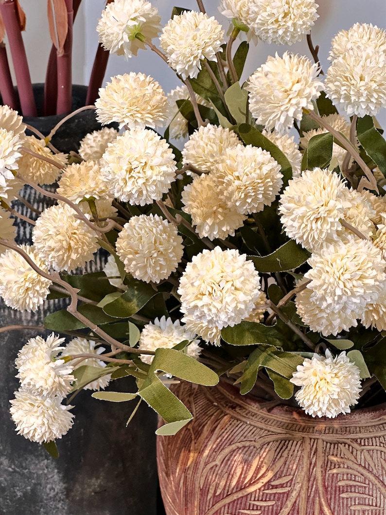 May include: A close-up of a floral arrangement featuring cream-colored, spherical flowers with green leaves. The flowers are arranged in a decorative, textured, brown vase. The arrangement is a natural, decorative element.