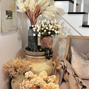May include: A decorative corner arrangement featuring various vases and floral arrangements. Tall, feathery beige and brown stems are in a dark vase, while smaller white ball-shaped flowers are in a brown vase. A gray chair with a throw blanket and pillow is on the right.