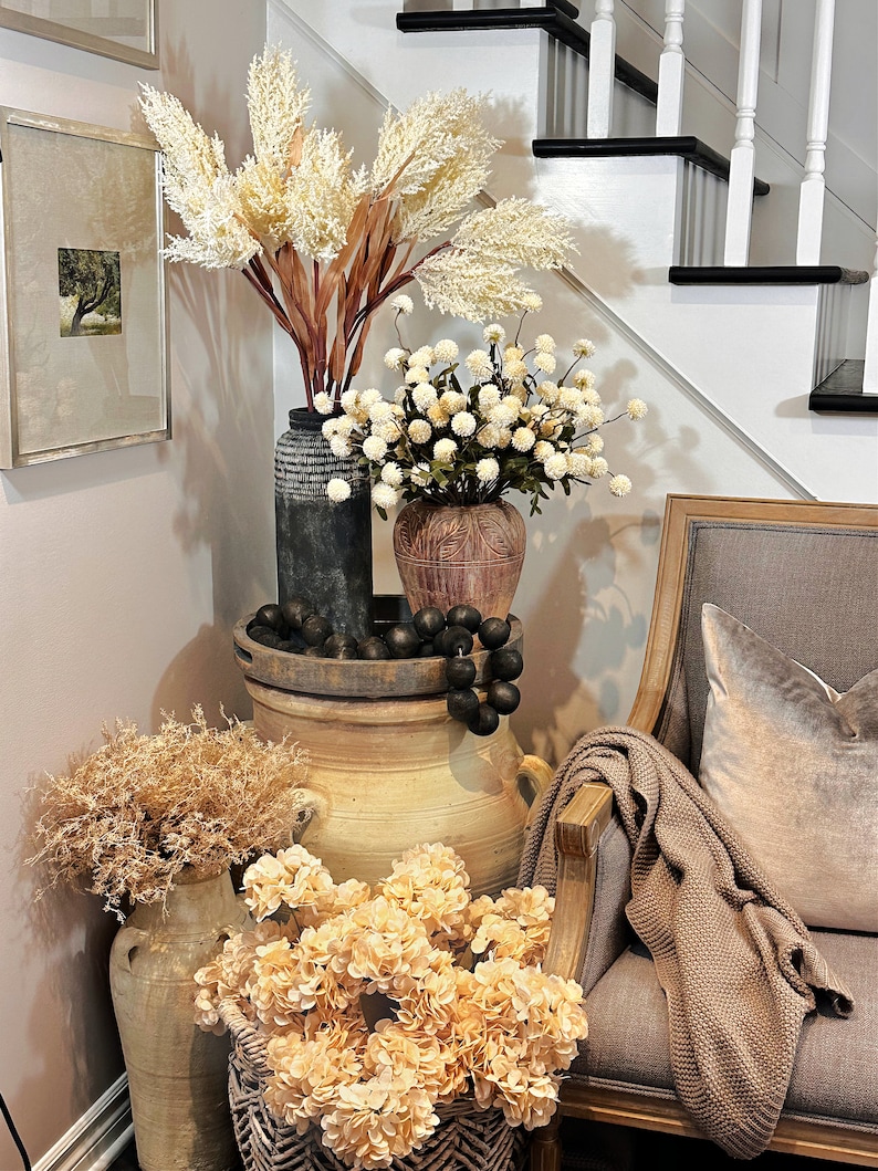 May include: A decorative corner arrangement featuring various vases and floral arrangements. Tall, feathery beige and brown stems are in a dark vase, while smaller white ball-shaped flowers are in a brown vase. A gray chair with a throw blanket and pillow is on the right.