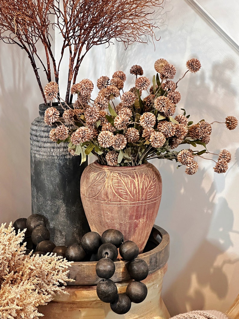 May include: A decorative arrangement featuring dried flowers and foliage in textured vases. The display includes a dark gray vase, a reddish-brown vase with carved details, and a wooden tray with dark wooden beads. The overall color palette is neutral with earthy tones.