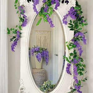 Op de afbeelding: Een ovale spiegel met wit frame, versierd met drie slingers van paarse blauwe regen en groene bladeren. De spiegel weerspiegelt een kamer met een macram&eacute; wandkleed en een vaas met blauwe regen. De tekst "Three garlands" staat bovenaan.