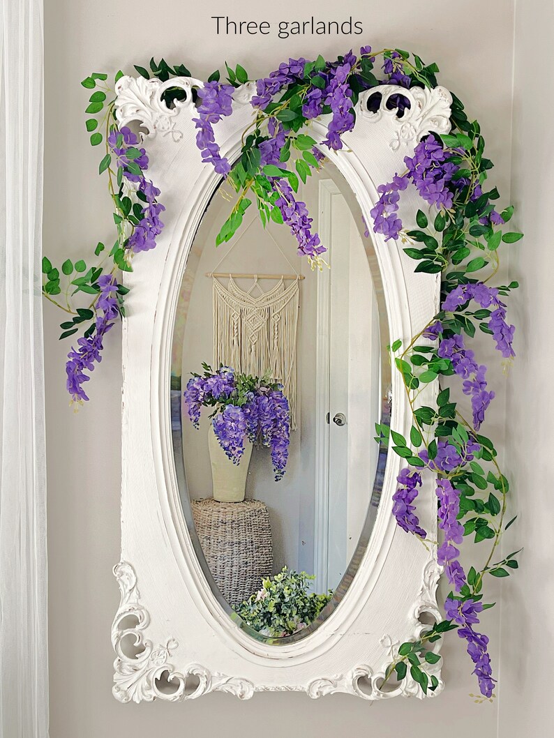 Op de afbeelding: Een ovale spiegel met wit frame, versierd met drie slingers van paarse blauwe regen en groene bladeren. De spiegel weerspiegelt een kamer met een macram&eacute; wandkleed en een vaas met blauwe regen. De tekst "Three garlands" staat bovenaan.