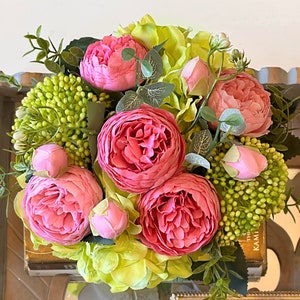 Op de afbeelding: Een boeket van roze pioenen en groene hortensia's in een vaas. De bloemen zijn in een cirkelvormige opstelling gerangschikt en omgeven door groene bladeren.