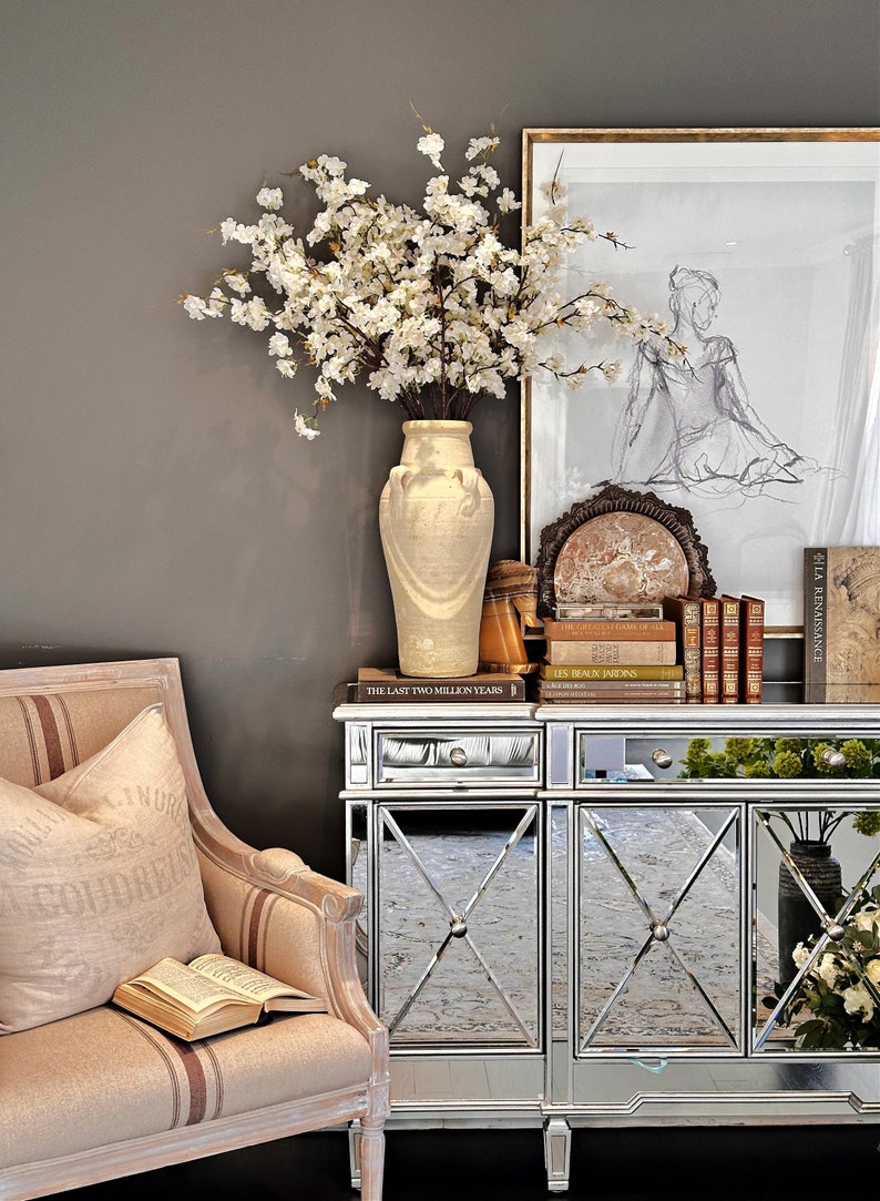 May include: A mirrored cabinet with a white ceramic vase filled with white blossoms. The cabinet has two drawers and two doors with a criss-cross pattern. The cabinet is sitting on a dark brown rug. The cabinet is decorated with books and a framed drawing of a woman.