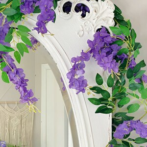 Op de afbeelding: Een witte, boogvormige spiegel is versierd met paarse wisteria en groene bladeren. De bovenkant van de spiegel heeft een verweerde, vintage uitstraling. Een macram&eacute; wandkleed is zichtbaar op de achtergrond, wat bijdraagt aan de decoratie.