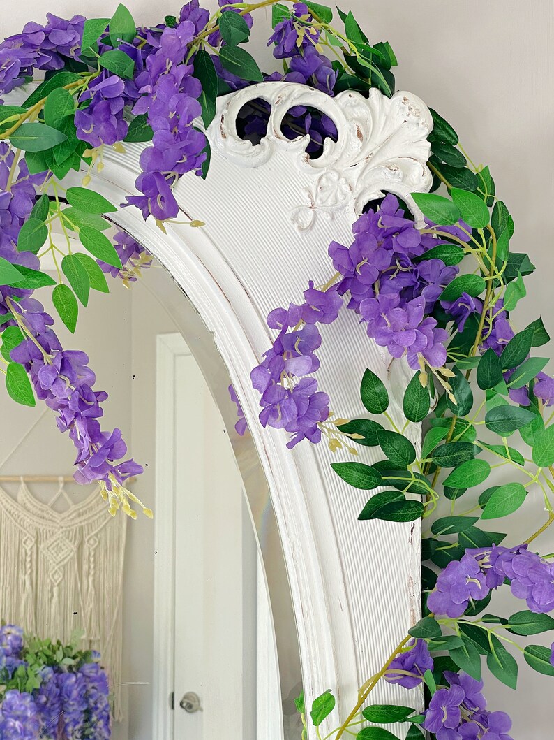 Op de afbeelding: Een witte, boogvormige spiegel is versierd met paarse wisteria en groene bladeren. De bovenkant van de spiegel heeft een verweerde, vintage uitstraling. Een macram&eacute; wandkleed is zichtbaar op de achtergrond, wat bijdraagt aan de decoratie.
