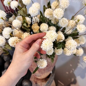 May include: Cream-colored artificial flower arrangement with spherical blooms and green leaves. The flowers are clustered together, creating a full and textured appearance. The arrangement is displayed in a wooden vase, suggesting a rustic or farmhouse aesthetic.
