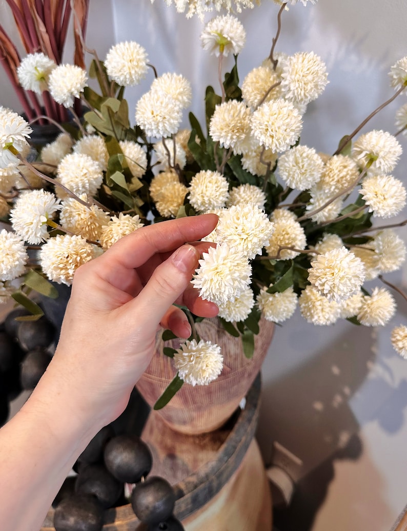 May include: Cream-colored artificial flower arrangement with spherical blooms and green leaves. The flowers are clustered together, creating a full and textured appearance. The arrangement is displayed in a wooden vase, suggesting a rustic or farmhouse aesthetic.