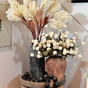 May include: A decorative arrangement featuring cream-colored faux pampas grass and white floral stems in textured vases. A wooden tray holds black wooden beads. The arrangement sits on a large, decorative ceramic pot.