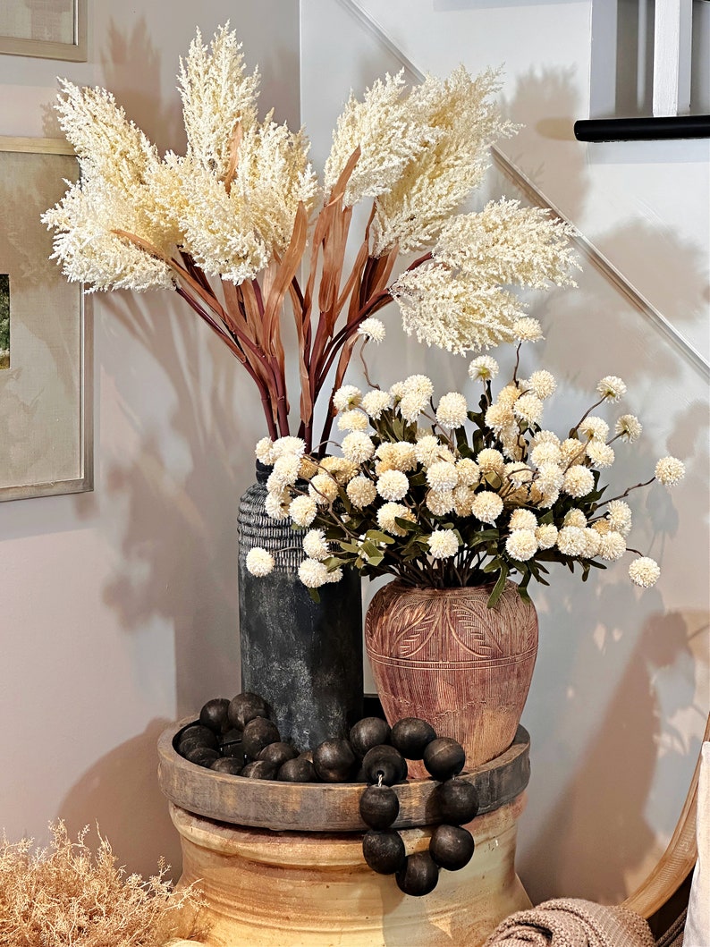 May include: A decorative arrangement featuring cream-colored faux pampas grass and white floral stems in textured vases. A wooden tray holds black wooden beads. The arrangement sits on a large, decorative ceramic pot.