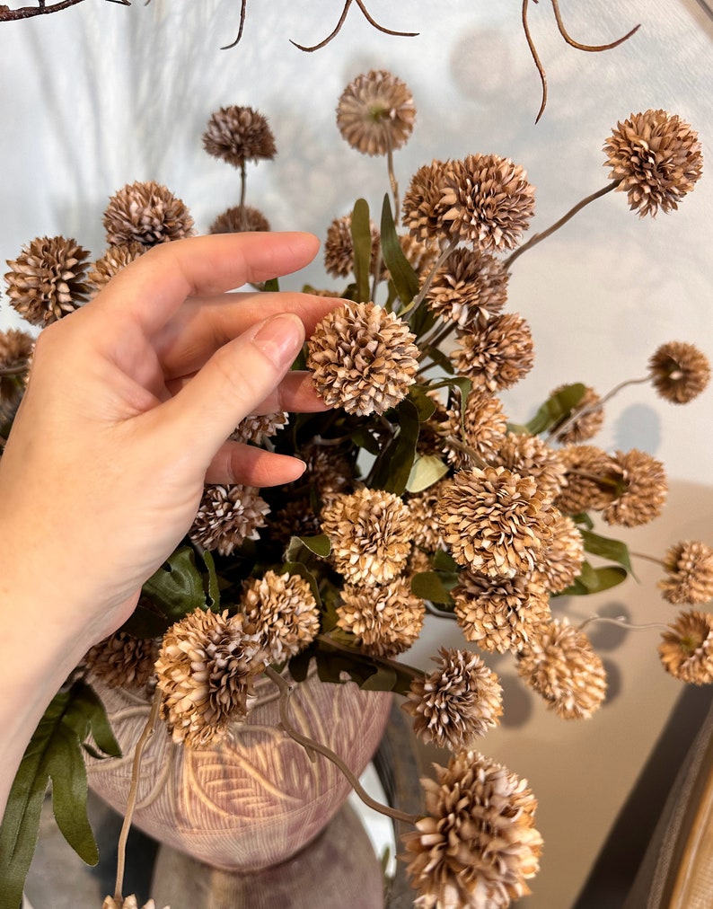May include: A bouquet of dried, spherical, tan-colored flowers with green leaves. The flowers are arranged in a decorative, textured vase. The arrangement is set against a neutral background, creating a natural, earthy aesthetic.