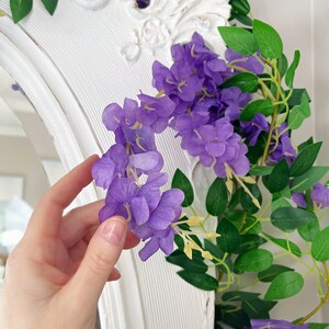 Op de afbeelding: Een close-up van een paarse wisteria slinger met groene bladeren. De slinger is gedrapeerd over een witte, sierlijke spiegel. De bloemen zijn levendig paars en de bladeren zijn diepgroen.