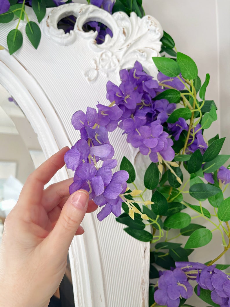 Op de afbeelding: Een close-up van een paarse wisteria slinger met groene bladeren. De slinger is gedrapeerd over een witte, sierlijke spiegel. De bloemen zijn levendig paars en de bladeren zijn diepgroen.