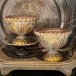 May include: Two decorative, antique-style bowls with a gold and teal finish. The bowls have a pedestal base and are adorned with floral detailing around the rim. They sit on a woven tray, with a large silver platter in the background.