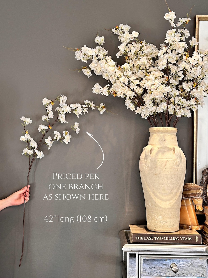 May include: A single branch of white cherry blossoms with a 42 inch (108 cm) length. The branch is held in a hand against a gray wall. The text "PRICED PER ONE BRANCH AS SHOWN HERE 42" long (108 cm)" is written below the branch.