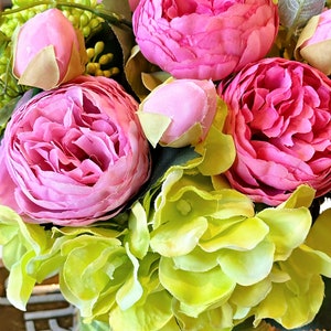 Op de afbeelding: Een boeket kunstbloemen in roze en groen in een groen-wit gestreepte vaas. De bloemen zijn in een tros gerangschikt en omvatten roze pioenen en groene hortensia's.
