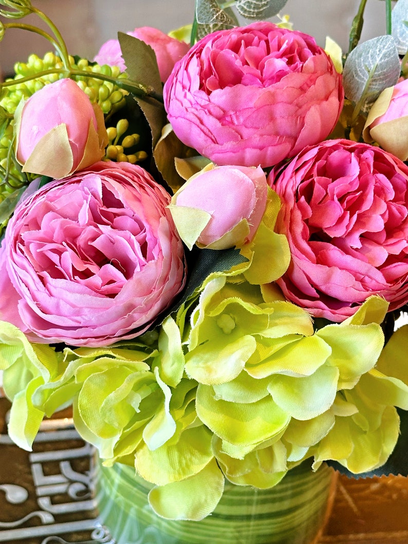 Op de afbeelding: Een boeket kunstbloemen in roze en groen in een groen-wit gestreepte vaas. De bloemen zijn in een tros gerangschikt en omvatten roze pioenen en groene hortensia's.