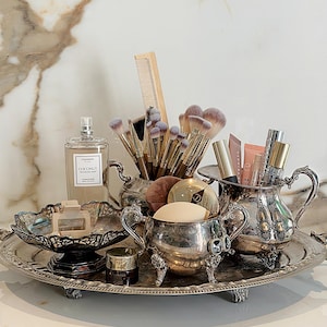 May include: A collection of silver-toned vintage vessels on an oval silver tray, holding makeup brushes, a wooden comb, fragrance mist labelled Coconut, lip products, and sponges. The set includes ornate bowls and a creamer pitcher with handles.