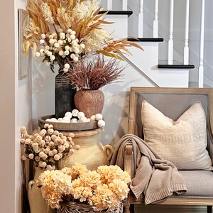 May include: A decorative arrangement featuring various vases and baskets filled with dried flowers and faux blooms in warm, autumnal tones. A gray armchair with a beige throw and pillow is to the right. A staircase is in the background.