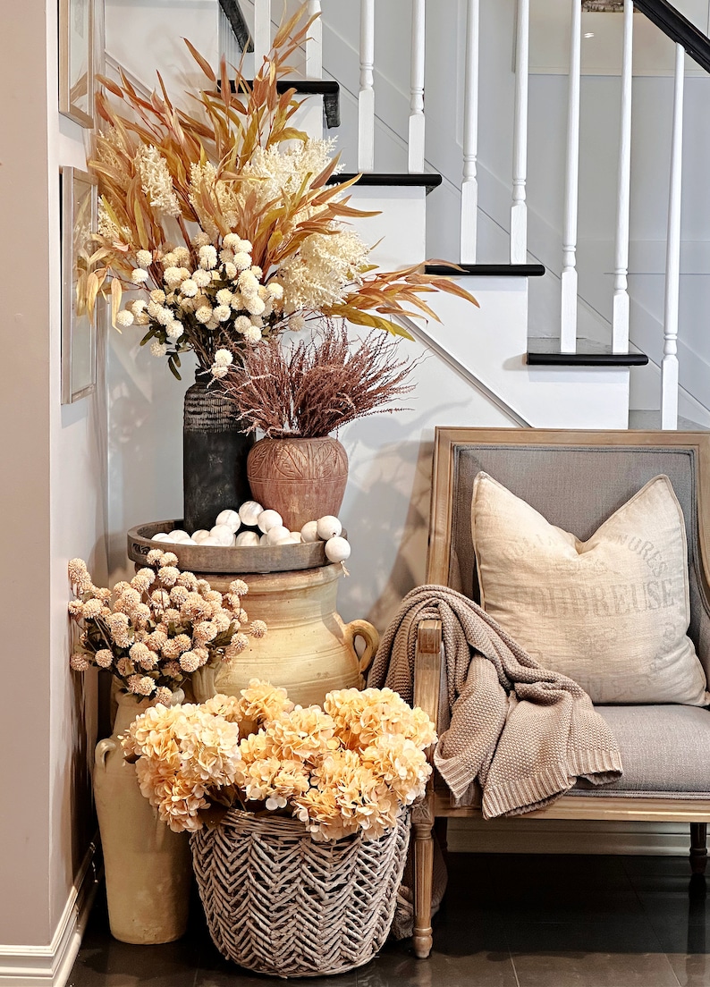 May include: A decorative arrangement featuring various vases and baskets filled with dried flowers and faux blooms in warm, autumnal tones. A gray armchair with a beige throw and pillow is to the right. A staircase is in the background.