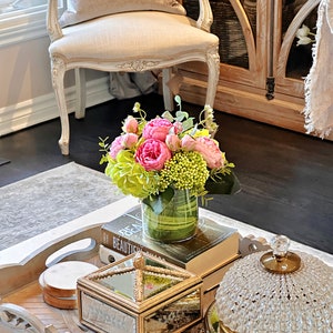 Op de afbeelding: Een houten dienblad met een decoratief ontwerp heeft een kristallen koepel, een spiegeldoos, een ronde houten onderzetter en een vaas met roze en groene bloemen. Het dienblad staat op een salontafel met een boek en een witte steen.